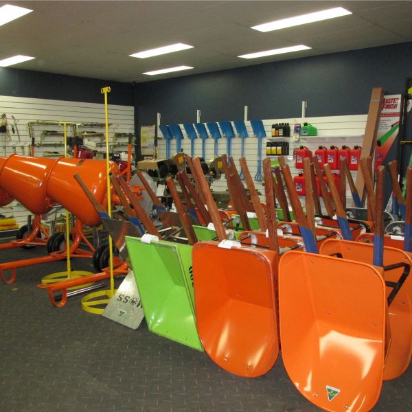 A hardware store showcasing various wheelbarrows in different colors, including orange and green, neatly lined up in front of the wall-mounted tool racks filled with construction tools and equipment.