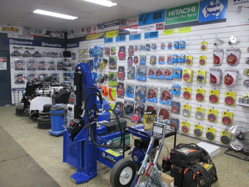 This image shows a display area in a tool shop, featuring a range of items on shelves, including products for cutting, grinding, and other construction tasks. In the foreground, there are various equipment pieces, such as a large blue tool with hoses, a vacuum cleaner, and other machinery. Several tool packages and parts are neatly arranged on the wall, with brands like Husqvarna and Hitachi visible.