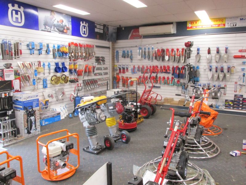 Various hand tools, such as trowels, hammers, and spanners, are neatly arranged on wall-mounted racks. Larger machinery items like a compactor and concrete finishing equipment are placed in the middle of the store, indicating a variety of tools for construction or industrial work. The shop is clean, with bright lighting and clear signage to help guide customers.